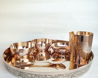 May include: A set of copper-toned bowls and a cup with intricate floral designs. The bowls and cup are arranged on a large, round copper-toned plate with a silver-toned border. The plate has a decorative floral design around the edge.