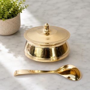 May include: A gold-colored, lidded bowl with a matching spoon. The bowl has a rounded shape with a lid and a small handle on top. The spoon is curved and lies next to the bowl. The items are on a marble surface.