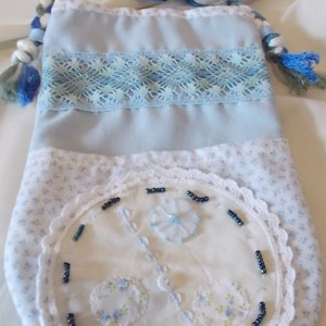 May include: A light blue handmade pouch with a braided strap in shades of blue and white. The pouch features a floral print, lace trim, and beaded embellishments. The design includes a circular applique with floral details.