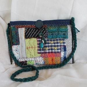 May include: A patchwork crossbody bag with a green, braided strap. The bag is made from various fabric squares in blue, green, black, and white, with decorative stitching and a button closure. A charm with turquoise beads hangs from the top.