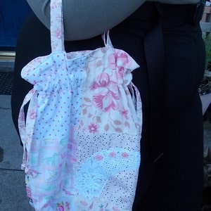 Op de afbeelding: Een patchwork-trekkoordtas met een bloemen- en stippenpatroon in roze en wit. De tas heeft een kanten en knoopdetail en een bijpassende bloemenband. De tas wordt door een persoon vastgehouden.