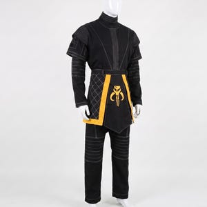 May include: A black long-sleeved tunic and pants set featuring white topstitching, a high collar, and short shoulder capes. A yellow waist sash with a quilted side panel and a central yellow Mythosaur skull emblem completes the ensemble.