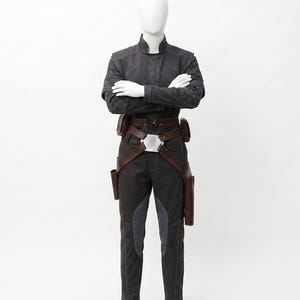 May include: A dark grey costume on a mannequin. The outfit features a jacket with diamond stitching on the shoulders, dark trousers with grey panels, and a brown belt with pouches and holsters. The mannequin's arms are crossed.