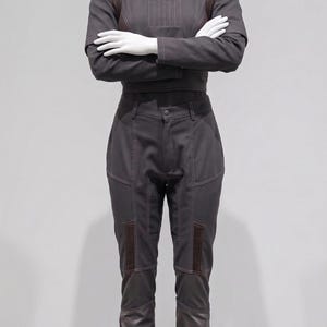 May include: A dark grey jumpsuit on a mannequin. The outfit has a high collar, long sleeves, and fitted trousers with brown accents. The stitching and paneling details enhance the design. The mannequin's arms are crossed.