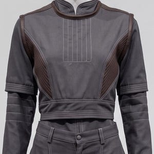 May include: A grey two-piece outfit with brown detailing and white stitching. The top features a high collar and quilted shoulder panels. The trousers have a button fastening and a tailored cut. The outfit is displayed on a mannequin.