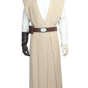 May include: A beige Jedi costume displayed on a mannequin. The costume includes a long tunic, a white shirt, and matching pants. A brown belt with a blue-green oval buckle and a black glove complete the outfit. The costume is designed for cosplay or themed events.