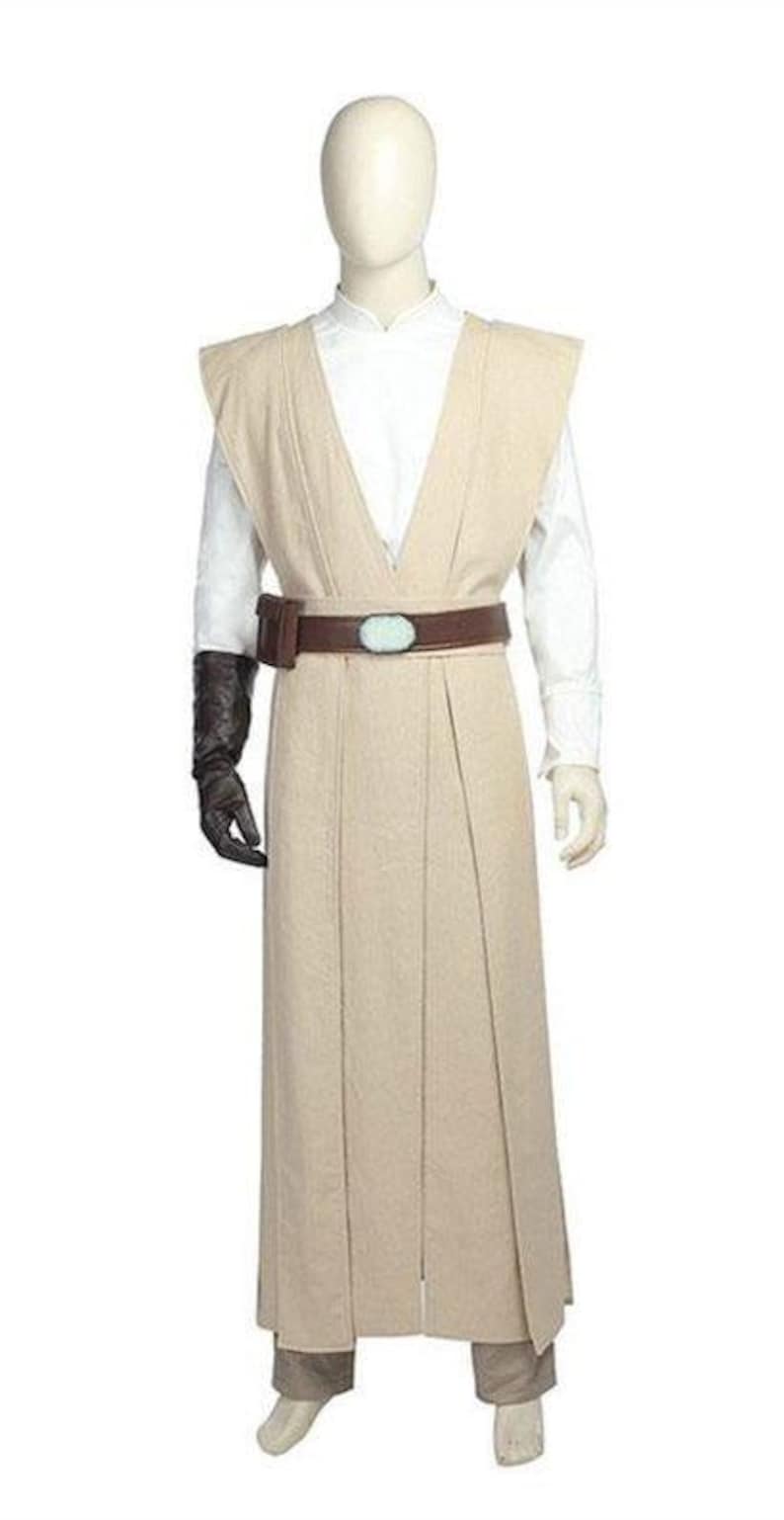 May include: A beige Jedi costume displayed on a mannequin. The costume includes a long tunic, a white shirt, and matching pants. A brown belt with a blue-green oval buckle and a black glove complete the outfit. The costume is designed for cosplay or themed events.