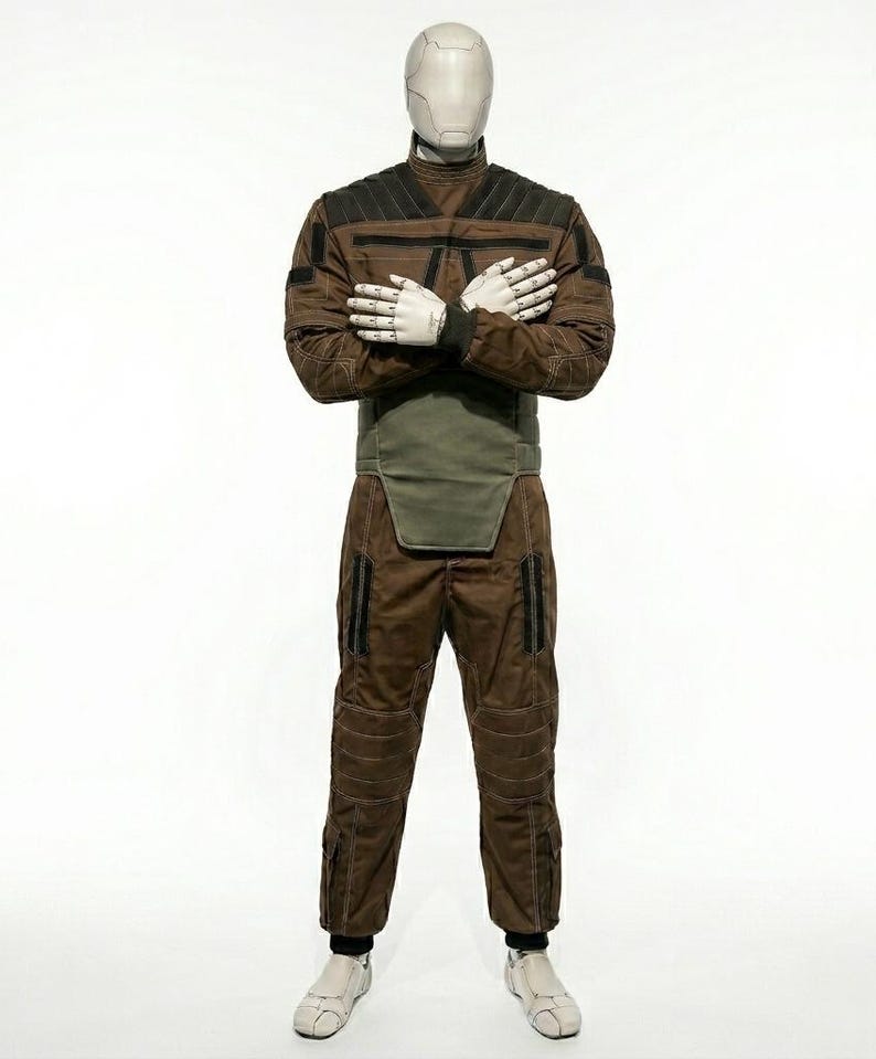 May include: A brown and black jumpsuit with a high collar, designed with quilted panels and pockets. The outfit includes a green waist protector and is displayed on a mannequin with a white head and hands. The mannequin is wearing white shoes.