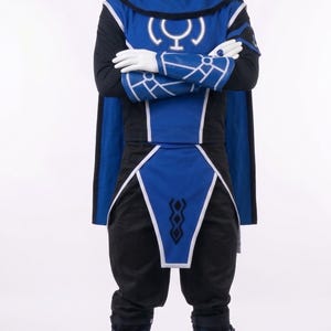 May include: A blue and black costume featuring a hooded tunic with a white circular symbol on the chest, matching arm gauntlets with white geometric patterns, a blue waist sash with a black diamond design, and blue boots with buckle straps.