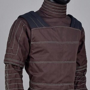 May include: A brown quilted garment with white stitching, featuring a high collar and shoulder pads. The design includes horizontal and vertical stitching patterns. The garment is displayed on a mannequin, showcasing its structured design.
