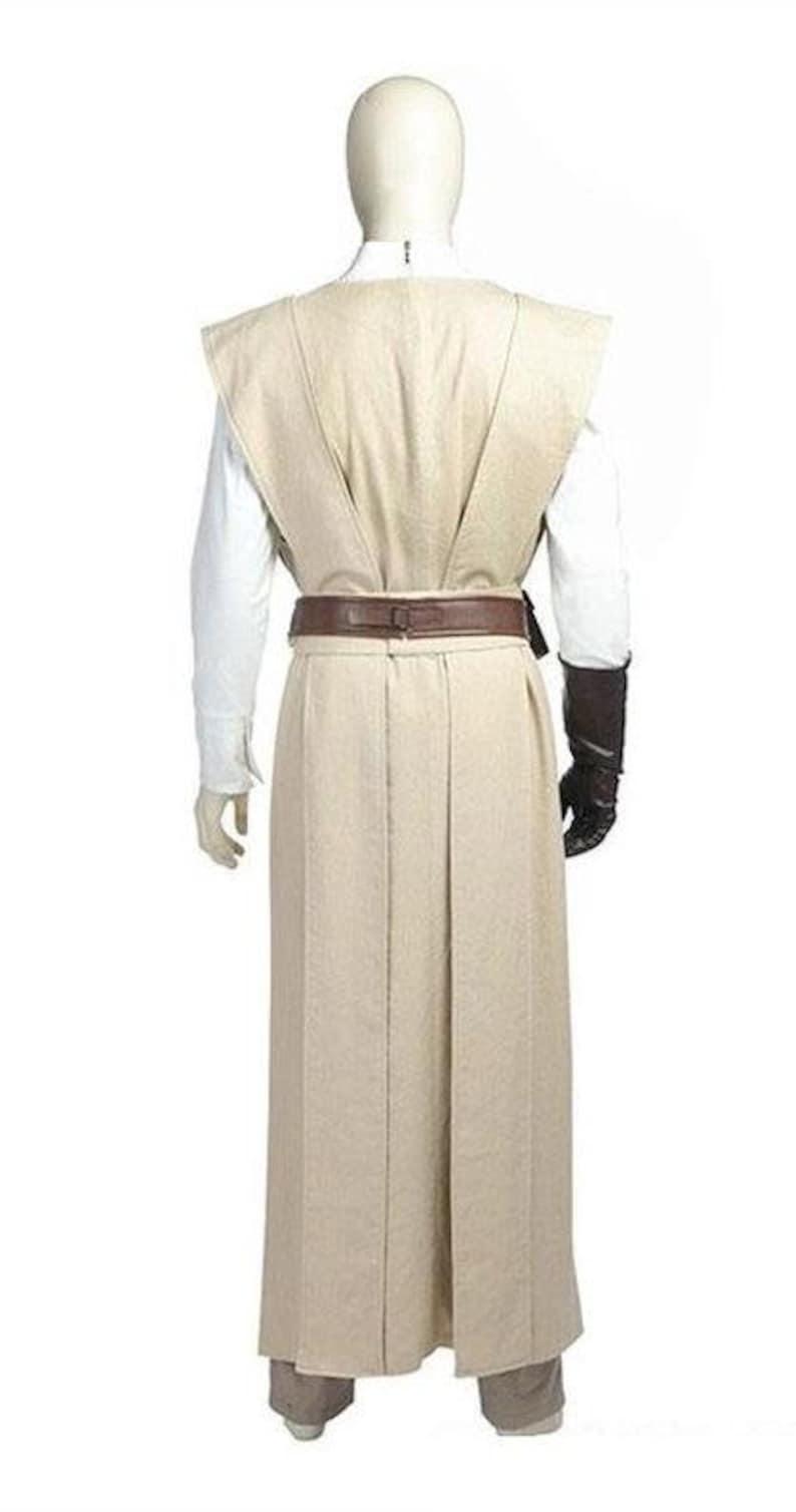 May include: A beige Jedi costume, featuring a long robe, a tabard, and a brown belt. The costume includes a white undershirt, brown gloves, and matching pants. The outfit is displayed on a mannequin against a white background.