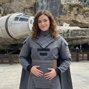 May include: A person wearing a gray Star Wars-themed outfit, including a long coat and pants, stands in front of the Millennium Falcon. The outfit has a brown belt and black accents. The background features rock formations and a cloudy sky.