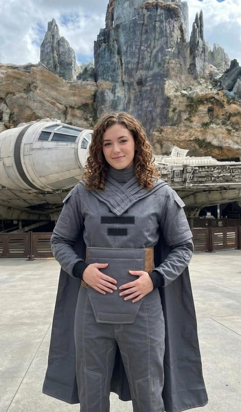 May include: A person wearing a gray Star Wars-themed outfit, including a long coat and pants, stands in front of the Millennium Falcon. The outfit has a brown belt and black accents. The background features rock formations and a cloudy sky.