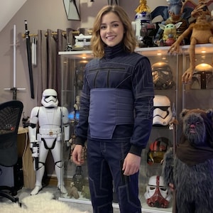 May include: A person wearing a dark blue jumpsuit with white stitching and a matching blue belt. The background includes Star Wars themed collectibles, including a TIE fighter model, Stormtrooper figure, and various character masks.