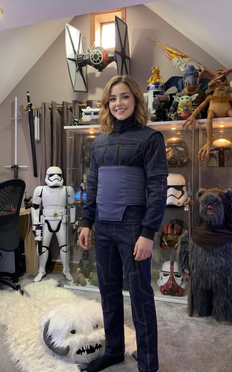 May include: A person wearing a dark blue jumpsuit with white stitching and a matching blue belt. The background includes Star Wars themed collectibles, including a TIE fighter model, Stormtrooper figure, and various character masks.