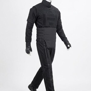 May include: A black tactical outfit displayed on a mannequin. The outfit includes a high-collared shirt, padded vest, belt, pants, gloves, and boots. White stitching accents the seams of the clothing. The mannequin is set against a white background.