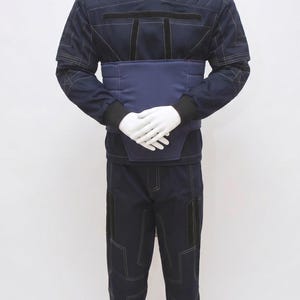 May include: Navy blue uniform displayed on a mannequin. The outfit features a high-collared jacket with quilted shoulders, a wide belt, and matching trousers. White stitching details the seams and pockets. The mannequin's hands are crossed at the waist.