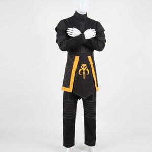 May include: A black long-sleeved tunic and matching trousers set featuring white topstitching details. The tunic includes a high collar and a yellow-trimmed apron-style panel with a yellow Mythosaur skull emblem centered on the front.