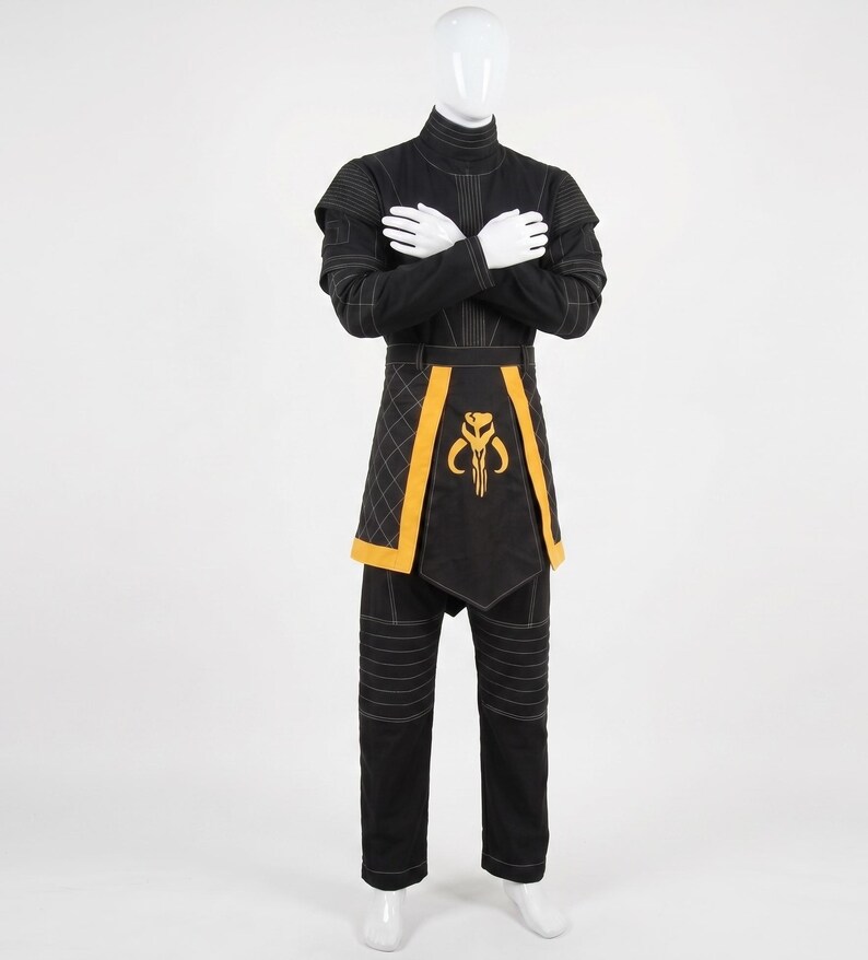 May include: A black long-sleeved tunic and matching trousers set featuring white topstitching details. The tunic includes a high collar and a yellow-trimmed apron-style panel with a yellow Mythosaur skull emblem centered on the front.
