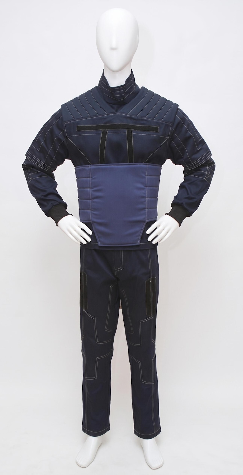 May include: A navy blue jumpsuit on a mannequin. The outfit has a high collar, quilted shoulder and chest panels, and a padded midsection. White stitching outlines the seams and pockets. The sleeves have black cuffs, and the trousers have geometric stitching.