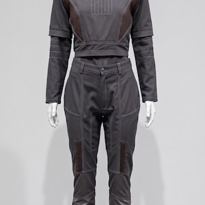 May include: A grey two-piece outfit displayed on a mannequin. The top features long sleeves and a high collar with brown detailing. The high-waisted trousers have brown patches on the knees and lower legs. The outfit is designed with visible stitching.