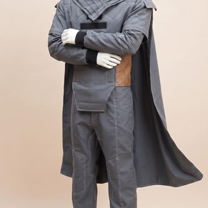 May include: A gray costume displayed on a mannequin. The outfit includes a jacket with a high collar, cape, and pants with white stitching. A brown belt is visible at the waist. The mannequin's arms are crossed.