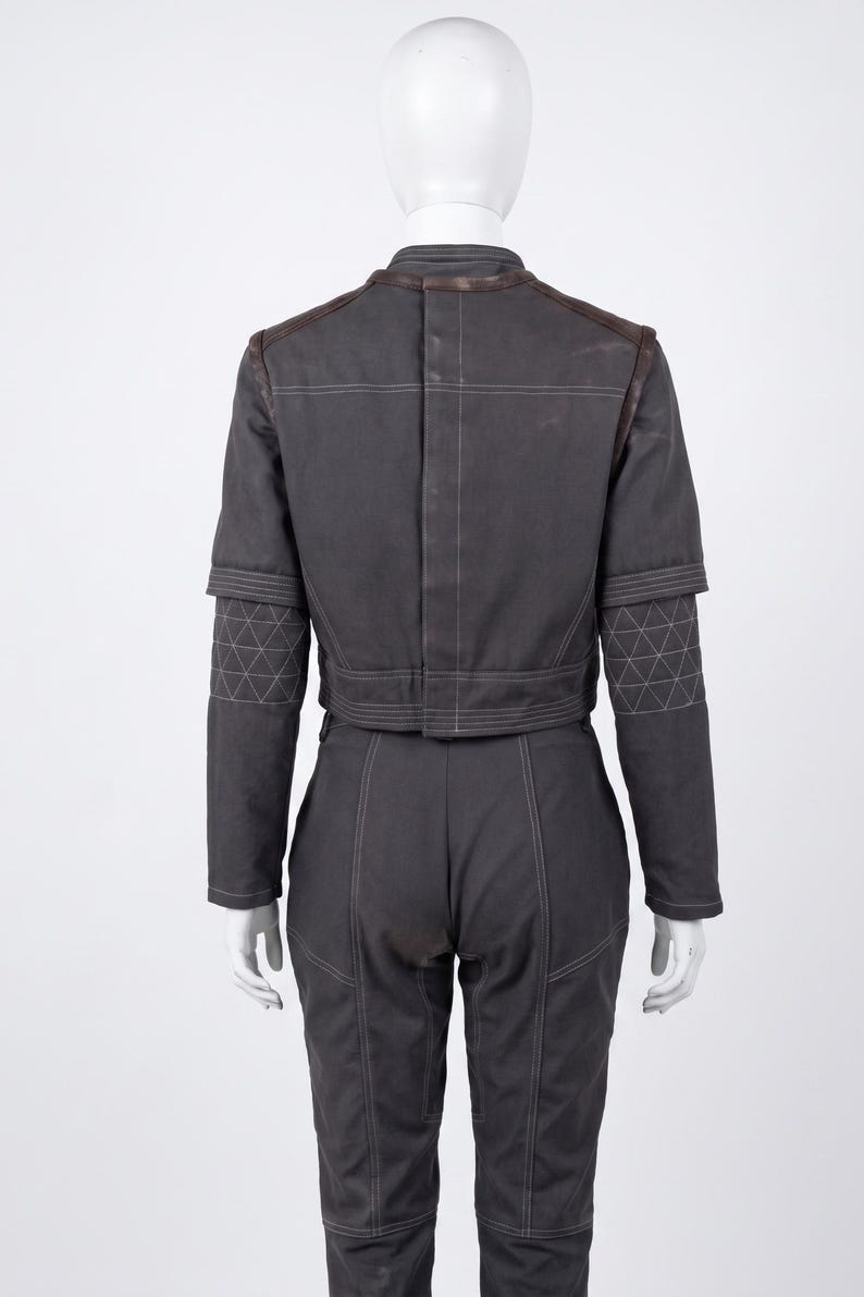 May include: Dark grey two-piece outfit. The jacket features brown leather detailing on the shoulders and collar, with geometric stitching on the sleeves. The trousers have matching stitching. The outfit is displayed on a mannequin.