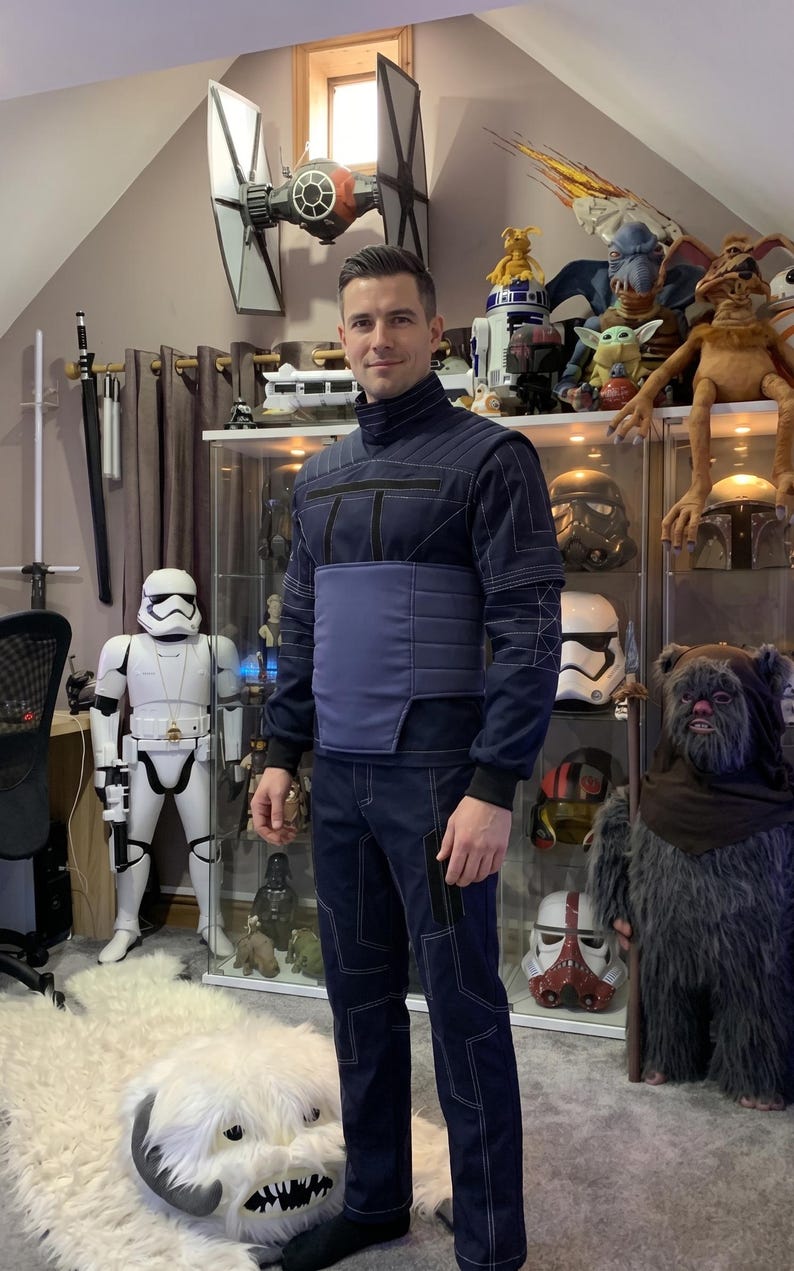 May include: A navy blue Star Wars-themed costume with white stitching and a matching vest. The costume is worn by a person standing in a room filled with Star Wars memorabilia, including a Stormtrooper figure and a TIE fighter model.
