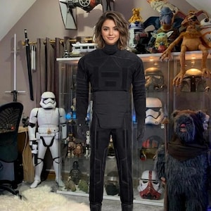 May include: A person wearing a black jumpsuit with a high collar, gloves, and boots, standing in a room filled with Star Wars memorabilia. A Stormtrooper figure and a Wampa rug are visible.