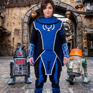 May include: A blue and black tunic with a white circular emblem on the chest, matching blue and black pants, and blue boots. The outfit is flanked by two small robotic droids, one dark gray with red accents and one beige with orange accents.