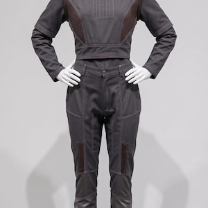 May include: A grey jumpsuit on a mannequin. The outfit has a high collar, long sleeves, and a cropped top with brown accents. The trousers are fitted with knee pads and the boots are dark brown.