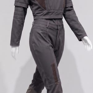 May include: A grey two-piece outfit on a mannequin. The top is a cropped jacket with brown accents and long sleeves. The trousers are fitted with brown patches on the knees. The outfit includes matching grey boots.