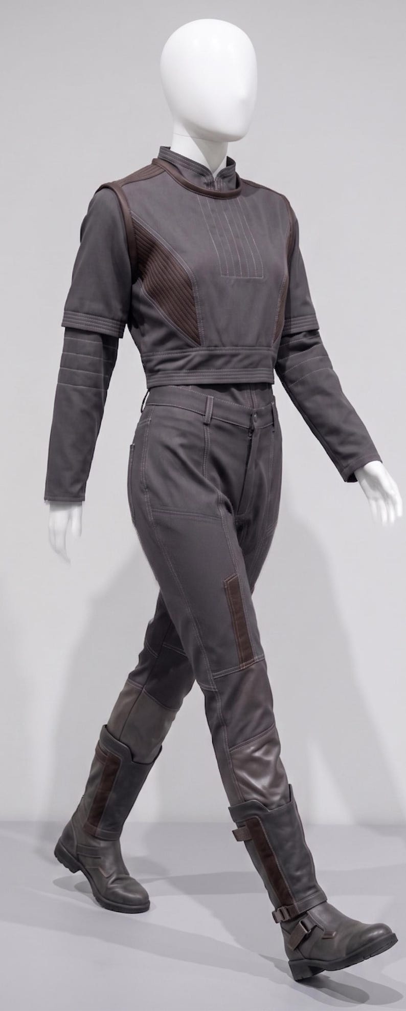 May include: A grey two-piece outfit on a mannequin. The top is a cropped jacket with brown accents and long sleeves. The trousers are fitted with brown patches on the knees. The outfit includes matching grey boots.
