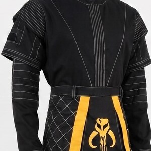 May include: A black long-sleeved tunic featuring a high collar, white topstitching, and layered shoulder panels. The lower section includes a black and yellow quilted apron-style panel with a yellow skull-shaped emblem centered on the front.