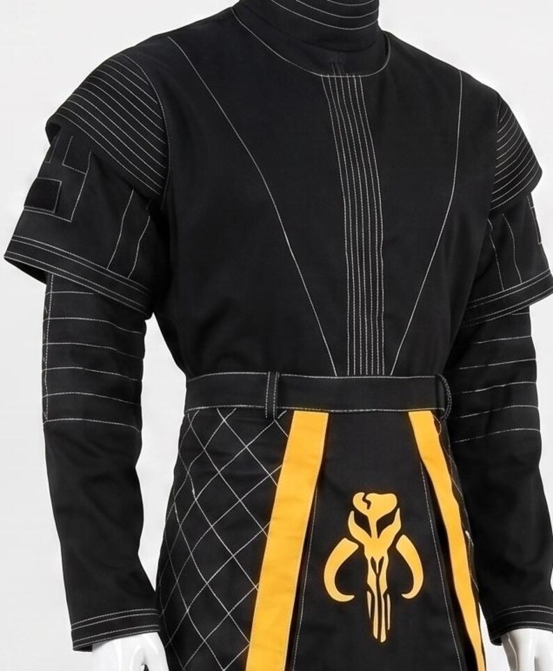 May include: A black long-sleeved tunic featuring a high collar, white topstitching, and layered shoulder panels. The lower section includes a black and yellow quilted apron-style panel with a yellow skull-shaped emblem centered on the front.