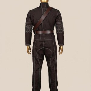 May include: A dark brown long-sleeved jacket and matching trousers set on a gold mannequin. The outfit features white contrast stitching, a high collar, quilted elbow patches, and a brown leather-like belt with a diagonal shoulder strap.