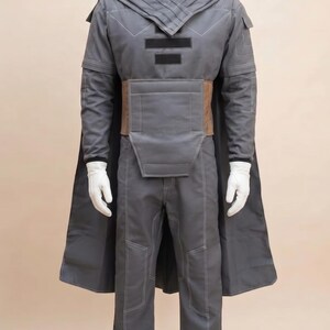 May include: A gray costume with a long cape, pants, and a jacket with a high collar. The jacket has a geometric design and a brown belt. The costume is displayed on a mannequin with white gloves.