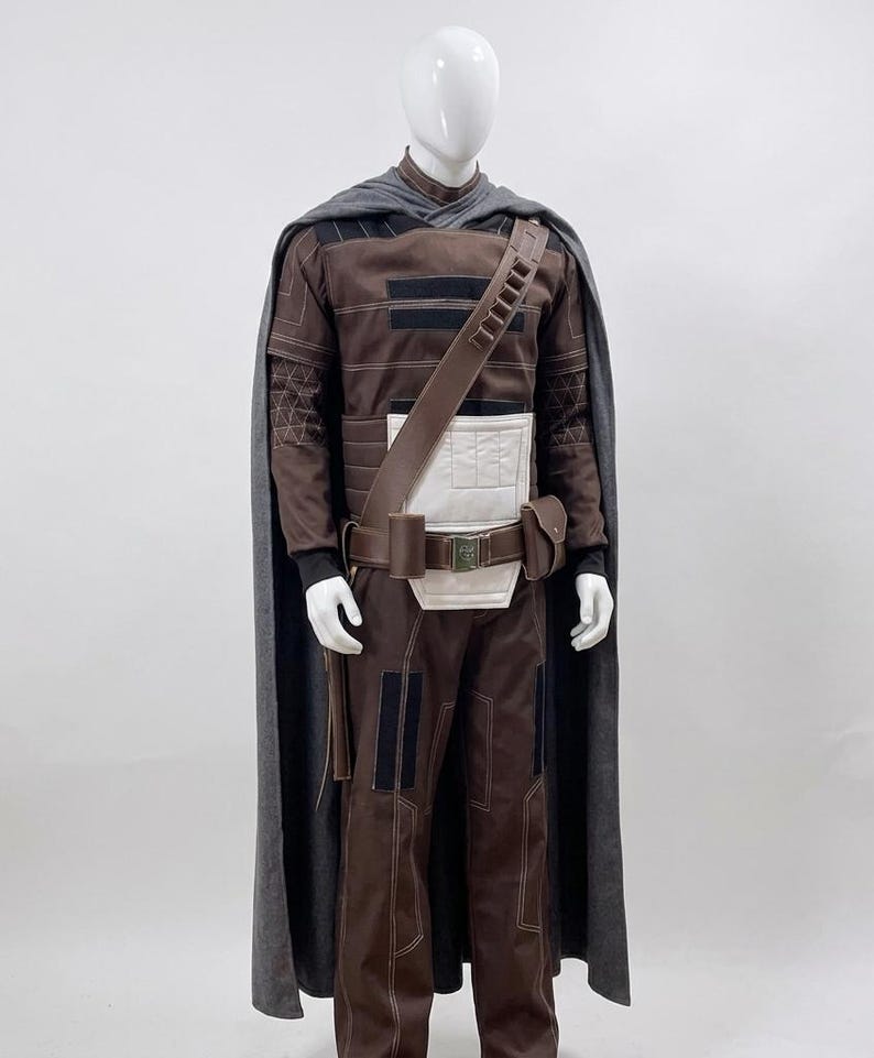 May include: A brown and black costume with a grey cape. The costume includes a long-sleeved shirt, trousers, and a belt with a pouch. The cape is draped over the shoulders. The costume is displayed on a mannequin.