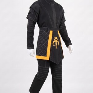 May include: A black costume set featuring a high-neck tunic with white topstitching, quilted side panels, and matching trousers. A yellow waist sash with a central yellow emblem featuring a stylized helmet design is worn over the tunic.