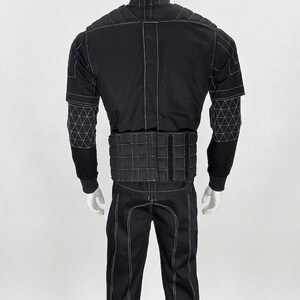 May include: Black jumpsuit with white stitching details, displayed on a mannequin. The outfit features a high collar, padded shoulders, and geometric patterns on the sleeves. The pants have a vertical seam down the front.