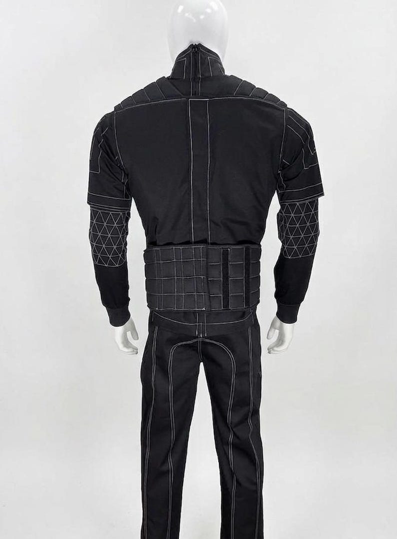 May include: Black jumpsuit with white stitching details, displayed on a mannequin. The outfit features a high collar, padded shoulders, and geometric patterns on the sleeves. The pants have a vertical seam down the front.