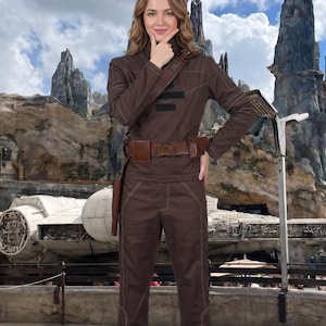 May include: A brown jumpsuit with white stitching, a brown belt, and matching boots. The outfit includes a holster and a shoulder strap. The background features a rock formation and a spaceship.