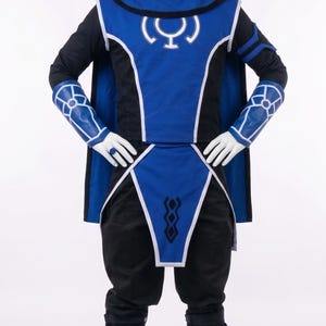 May include: A blue and black costume featuring a hooded tunic with a white circular emblem on the chest, matching arm guards with white geometric patterns, a long cape, black trousers, and blue boots with straps, displayed on a white mannequin.