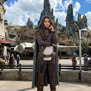 May include: A person in a brown and grey costume with a hooded cape, brown trousers, and boots. The costume includes a white chest piece and a utility belt. The background shows a themed environment with rock formations and a spaceship.