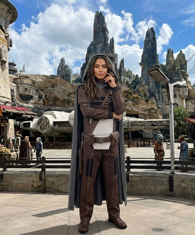 May include: A person in a brown and grey costume with a hooded cape, brown trousers, and boots. The costume includes a white chest piece and a utility belt. The background shows a themed environment with rock formations and a spaceship.