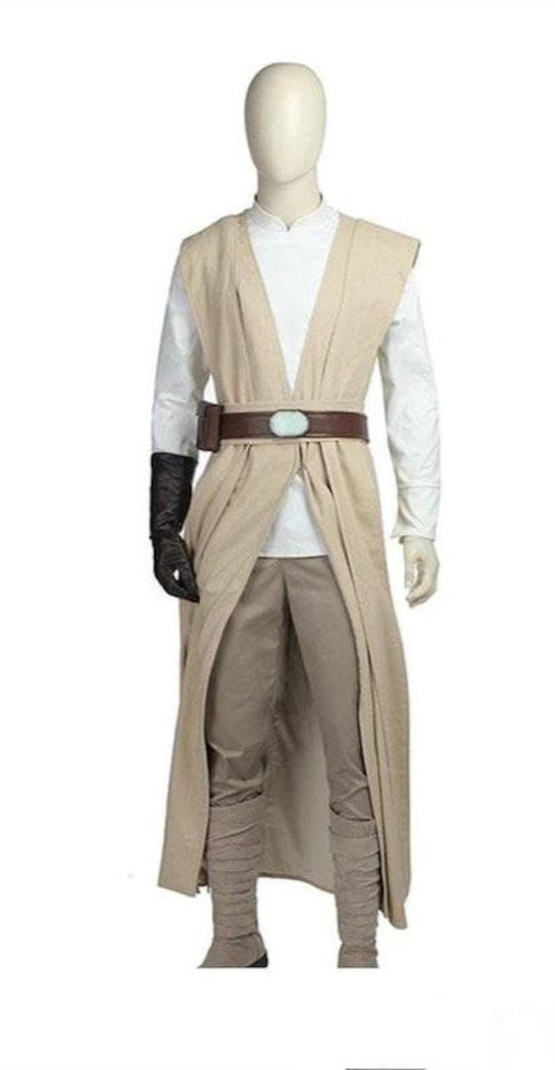 May include: A beige Jedi costume featuring a long, open-front robe, a white tunic, and matching pants. The costume includes a brown belt with a light blue buckle, a black glove, and matching boots. The outfit is displayed on a mannequin.