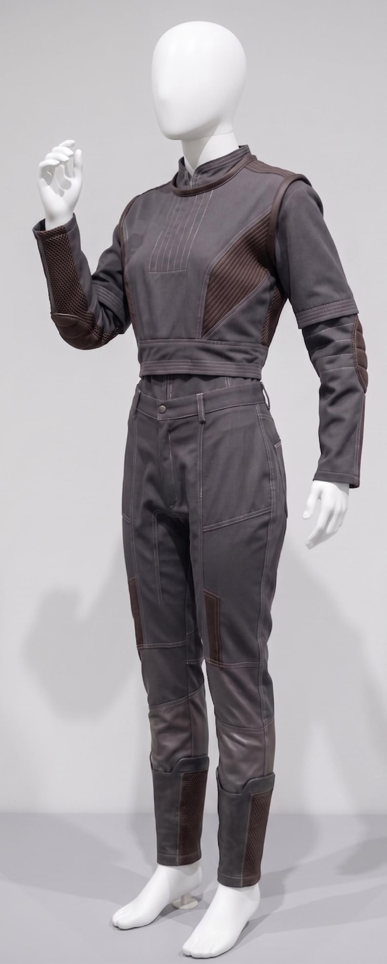 May include: A grey jumpsuit on a mannequin. The outfit includes a cropped top with brown detailing on the shoulders and sleeves, and matching trousers. The design features stitched details and reinforced patches.
