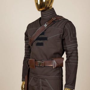 May include: A dark brown long-sleeved tactical outfit featuring a high collar, white topstitching, and a brown utility belt with pouches. A brown shoulder strap with a buckle crosses the chest, which displays three horizontal black rectangular patches.