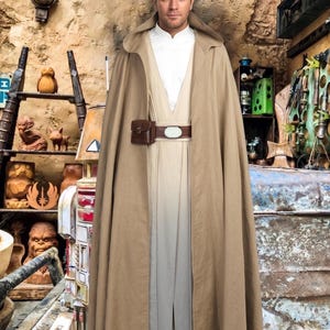 May include: A person wearing a tan cloak and tunic, with a brown belt and pouch. The outfit is reminiscent of a character from a science fiction film. The background features a rustic, themed environment with various props.
