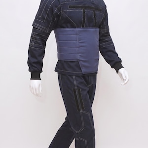 May include: A navy blue jumpsuit on a mannequin. The outfit has a high collar, long sleeves, and a contrasting blue panel across the midsection. White stitching details the seams. The trousers are straight-legged.
